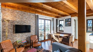 A spacious living area with a stone wall, wooden ceiling beams, a flat-screen TV, leather furniture, and a view to the outside. - Chalets am Diemelsee in Heringhausen (Diemelsee)