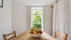 A wooden dining table with a bowl of fruit in the center, two wooden chairs, a window with a view of greenery, and a white curtain. - Gorgeous Garden Flat - Top Location (Cotham)