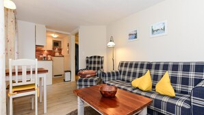 A cozy living room with a plaid sofa, a wooden table, and a white chair. A kitchen is visible in the background. - Ferienwohnung in Wyk-boldixum auf Foehr (Wyk auf Foehr)