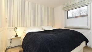 A bedroom with a bed, a nightstand, a lamp, a window with blinds, and a striped wall. - Ferienwohnung in Wyk-boldixum auf Foehr (Wyk auf Foehr)