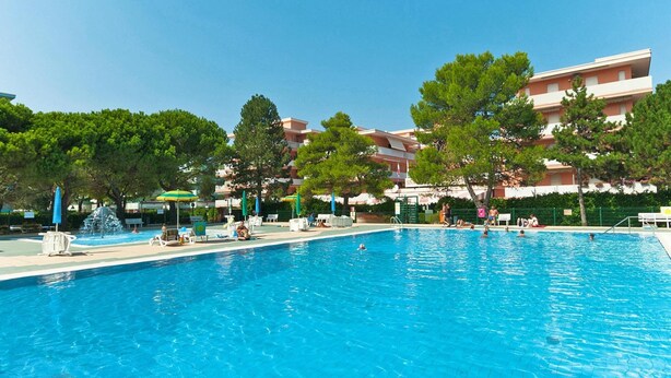 A swimming pool with lounge chairs and umbrellas, surrounded by trees and buildings. - Residence Valbella Beach (San Michele al Tagliamento)