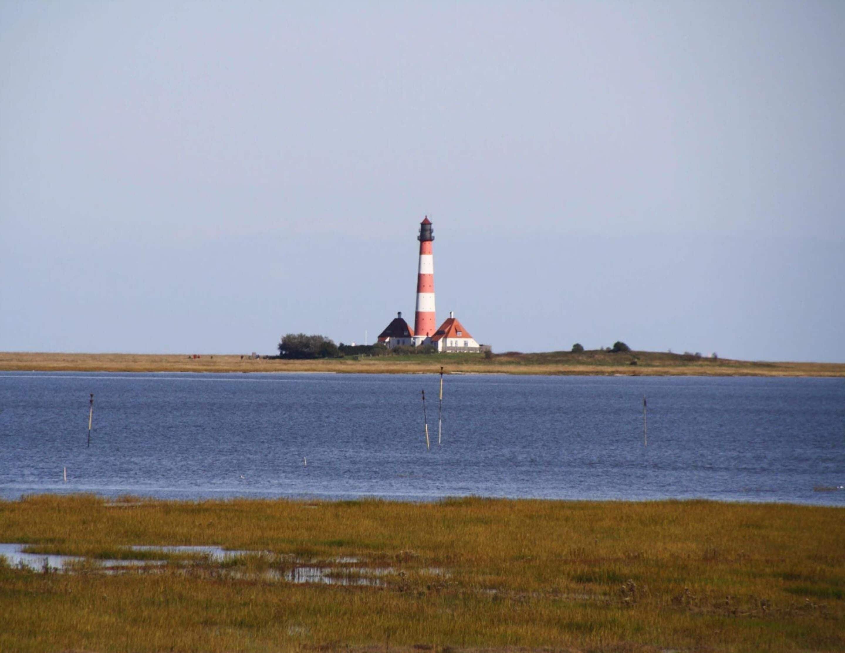 Un faro con una torre a rayas roja y blanca, un pequeño edificio blanco de techo rojo y una orilla cubierta de césped con agua en calma en primer plano.