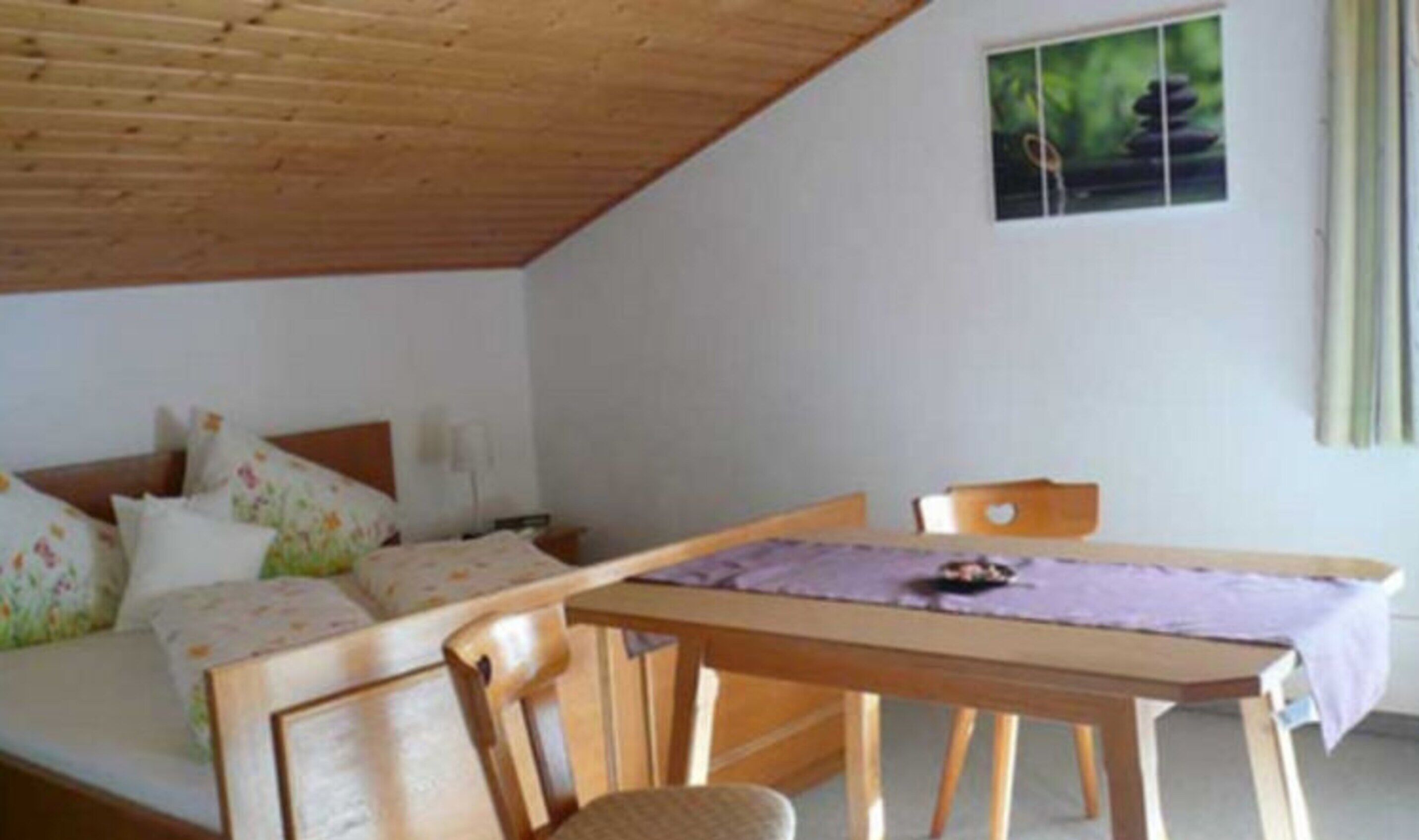 Una camera accogliente con soffitto in legno, un letto con biancheria da letto floreale, un tavolo con tovaglia viola e una finestra vista su vegetazione.