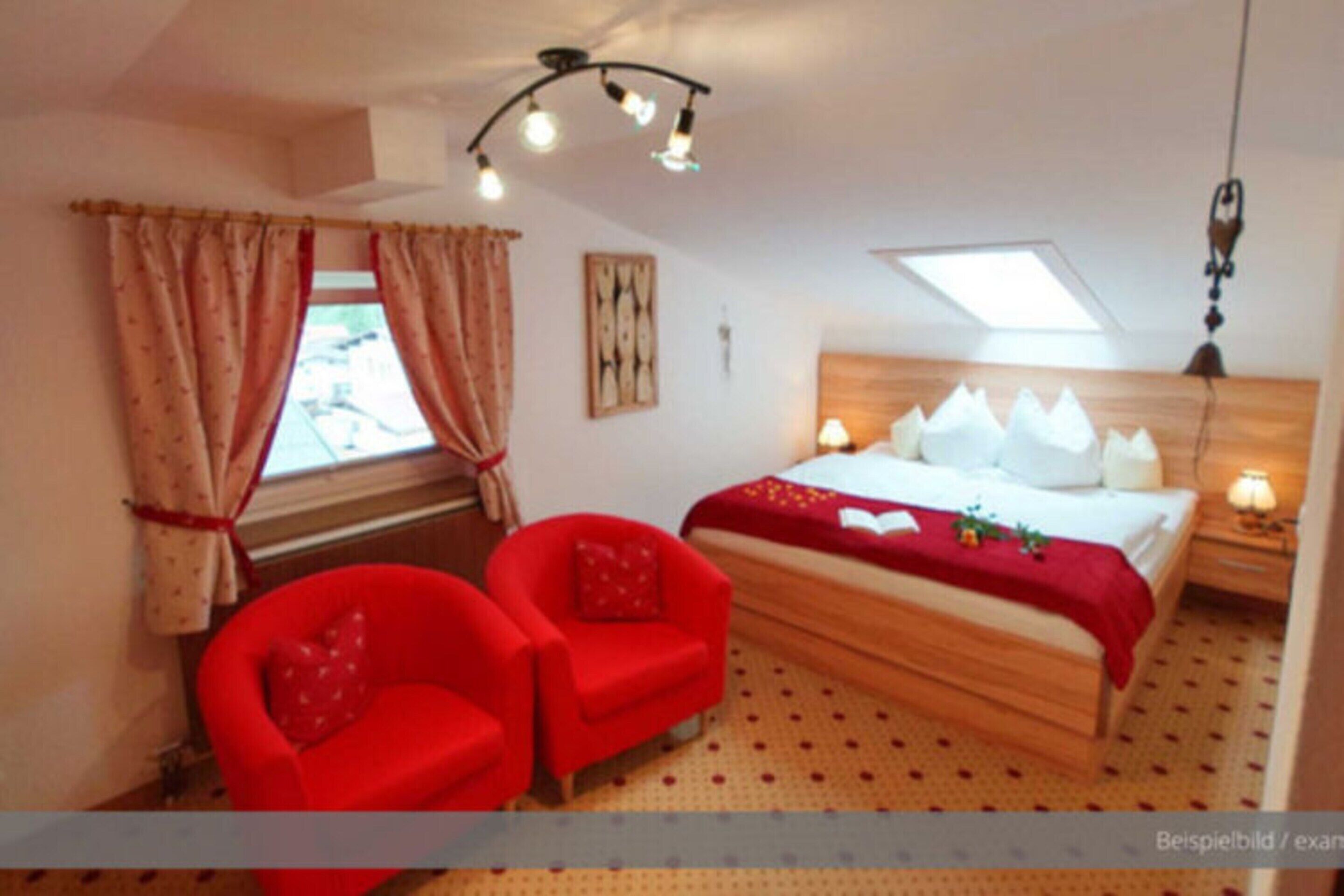 Ein Schlafzimmer mit einem Bett, zwei roten Sesseln, einem Fenster mit Vorhängen, einer Deckenleuchte und einem an der Wand hängenden Kunstwerk.