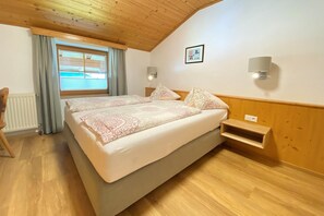 Een slaapkamer met een groot bed, houten plafond, een raam met gordijnen, een nachtkastje en een wandlamp.
