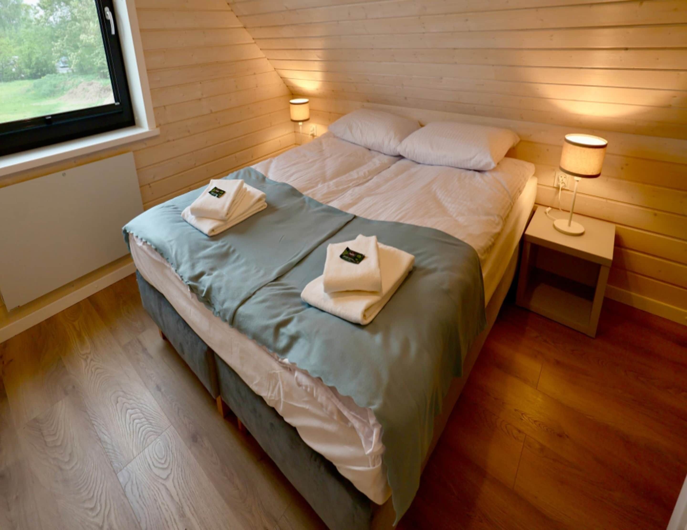 A neatly made bed with two folded towels, a bedside lamp, and a window with a view of greenery.