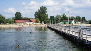 Ein Schwimmbereich an einem Fluss mit einer Holzsteganlage, einem Sandstrand und Gebäuden im Hintergrund.