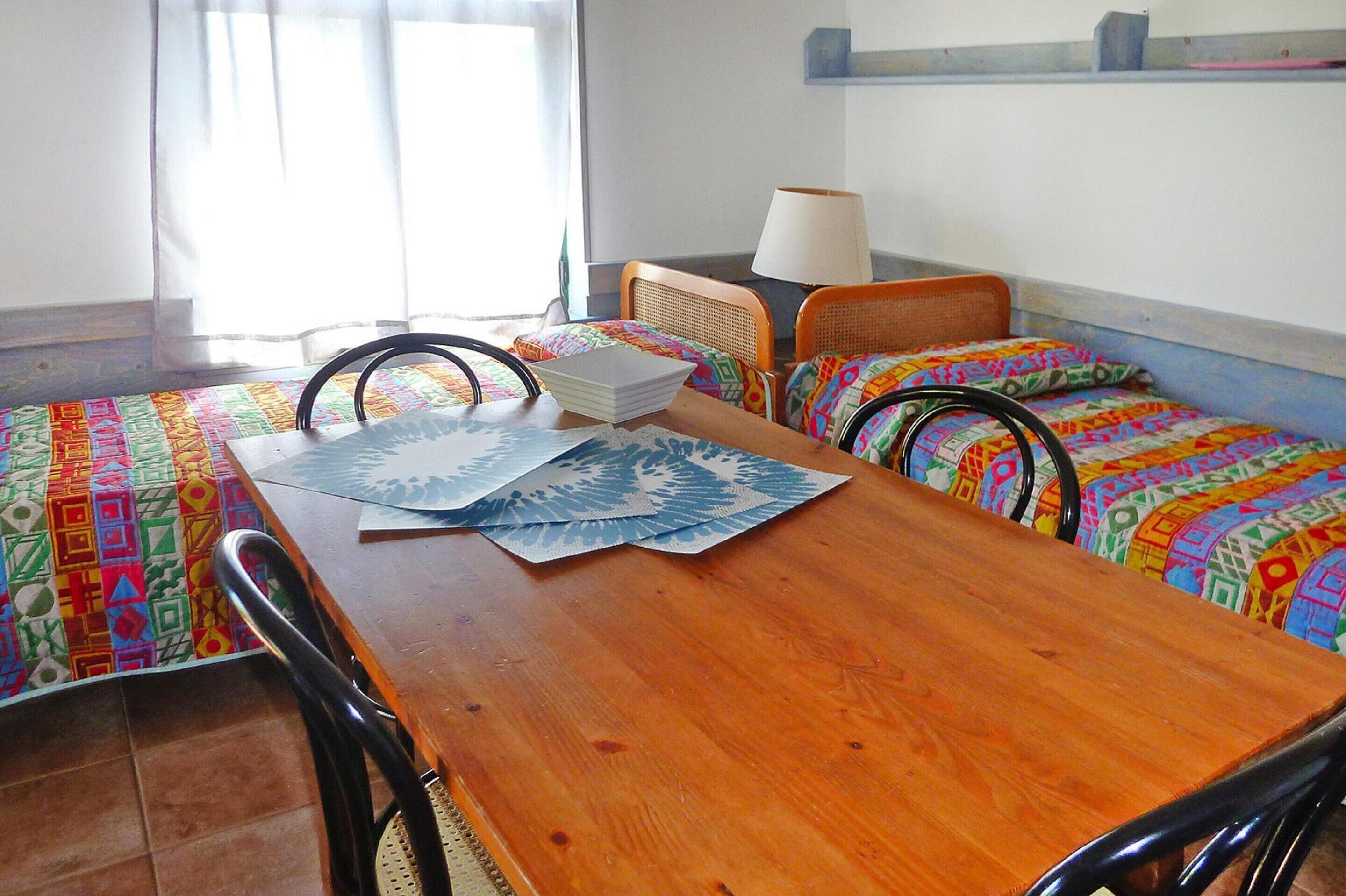 A room with a wooden table, two beds with colorful bedding, a chair, a lamp, and a window with curtains.