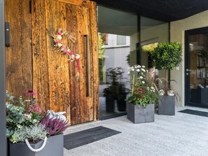 Una puerta de madera con un arreglo floral, plantas en macetas y la entrada a un edificio moderno.