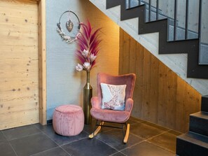 Un intérieur moderne avec un mur en bois, un fauteuil rose recouvert de tissu, agrémenté d’un coussin décoratif, un petit pouf rond et un vase contenant des fleurs séchées.