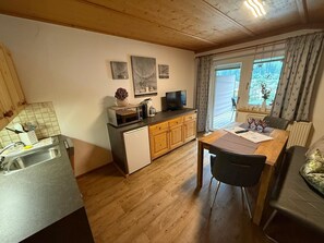 Una cocina compacta con microondas, cafetera y refrigerador pequeño. Hay una zona de comedor con mesa y sillas, y una ventana con cortinas.