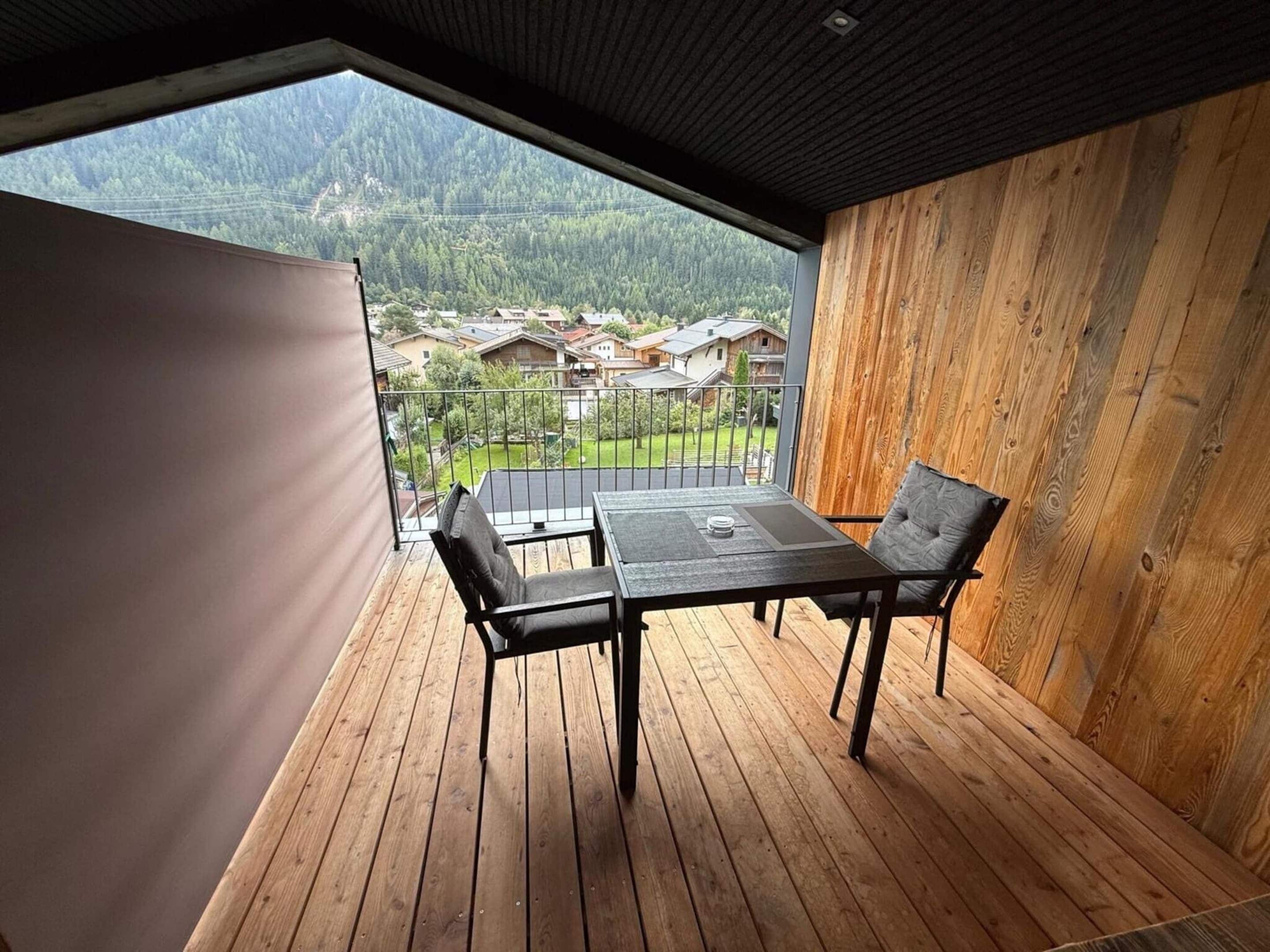 A balcony with a table and chairs, wooden flooring, and a view of a residential area and mountains.