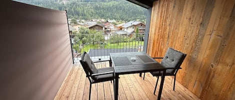 A balcony with a table and chairs, wooden flooring, and a view of a residential area and mountains.
