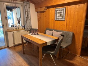 Une table à manger en bois, recouverte d’un chemin de table blanc, avec deux chaises grises à coussin et trois verres en cristal posés sur la table.