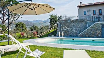 A swimming pool with a stone wall and a staircase leading to a building. There are lounge chairs and an umbrella.