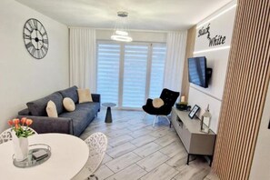 A modern living room with a grey sofa, a round white table, and a black chair. There is a TV mounted on the wall and a window with white curtains. - Apartment With a Terrace in Mielno (Mielno)