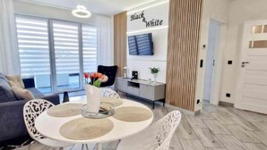 Un salon moderne avec une table à manger ronde, des chaises blanches et un canapé. Un téléviseur à écran plat est fixé au mur, et un panneau décoratif porte l’inscription « Noir et blanc ».