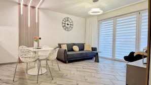 Une cuisine moderne avec des meubles blancs, une table à manger avec des chaises blanches, un canapé gris avec des coussins et une grande fenêtre avec des stores.