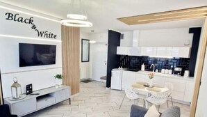 Un salon moderne avec un canapé gris, une petite table ronde et un téléviseur fixé au mur. On y trouve également un coin cuisine avec des meubles blancs et un plan de travail noir.