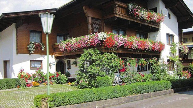 En traditionell alpchata med en balkong som är prydd med färgstarka blommor, en välskött trädgård och en klar himmel.