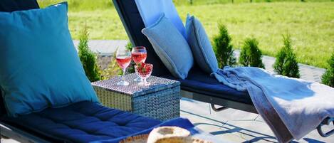 A sun lounger with a cushion, a small table with glasses and a basket, and a blanket on a patio.