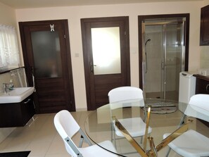 A compact room with a glass table surrounded by white chairs, a sink, and a shower area.