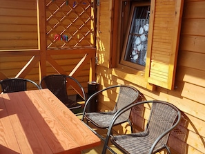 Eine Holzterrasse mit Tisch und Stühlen, ein Fenster mit Fensterläden und eine Gittertür.