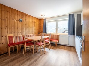 Une pièce aux murs lambrés, comprenant une table à manger, des chaises, une horloge, une fenêtre avec des rideaux et un radiateur.