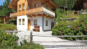 A two-story wooden chalet with a balcony, surrounded by lush greenery and a stone pathway.