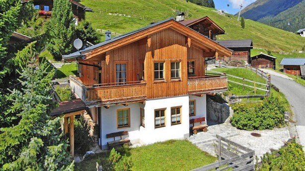 A wooden chalet with a balcony, surrounded by lush greenery and mountains.