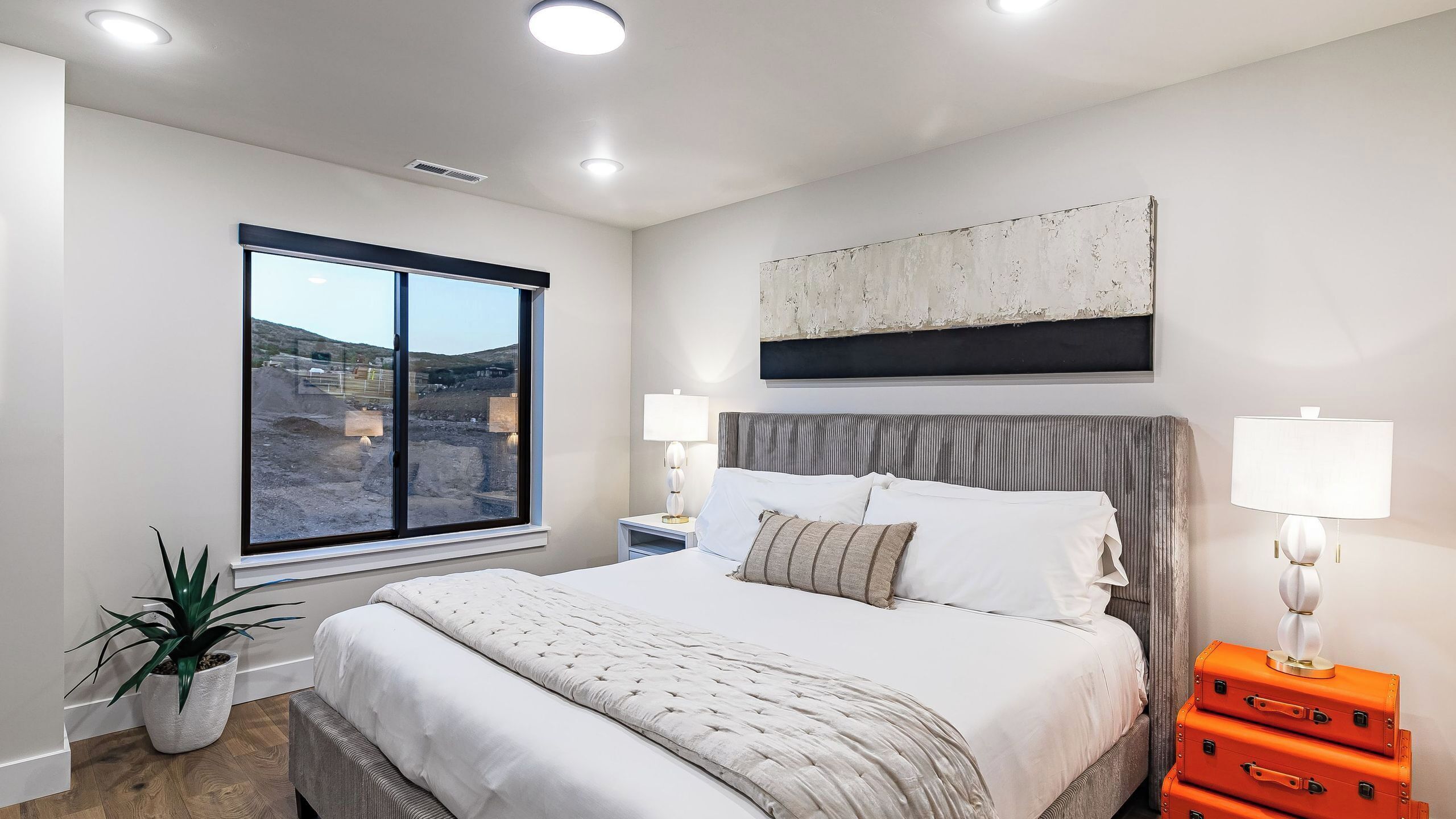 A bedroom with a large bed, two bedside tables with lamps, a window with a view of a construction site, and a potted plant.
