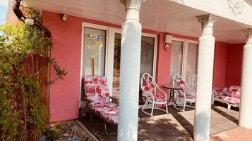 A covered patio with white chairs and a small table, a red wall, and a window with a closed shutter.