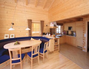 A cozy cabin interior with a wooden dining table, blue cushioned seating, a kitchen area with wooden cabinets, and a large window with plaid curtains. - Chalet in Konigsleiten Near Hohe Tauern (Wald im Pinzgau)