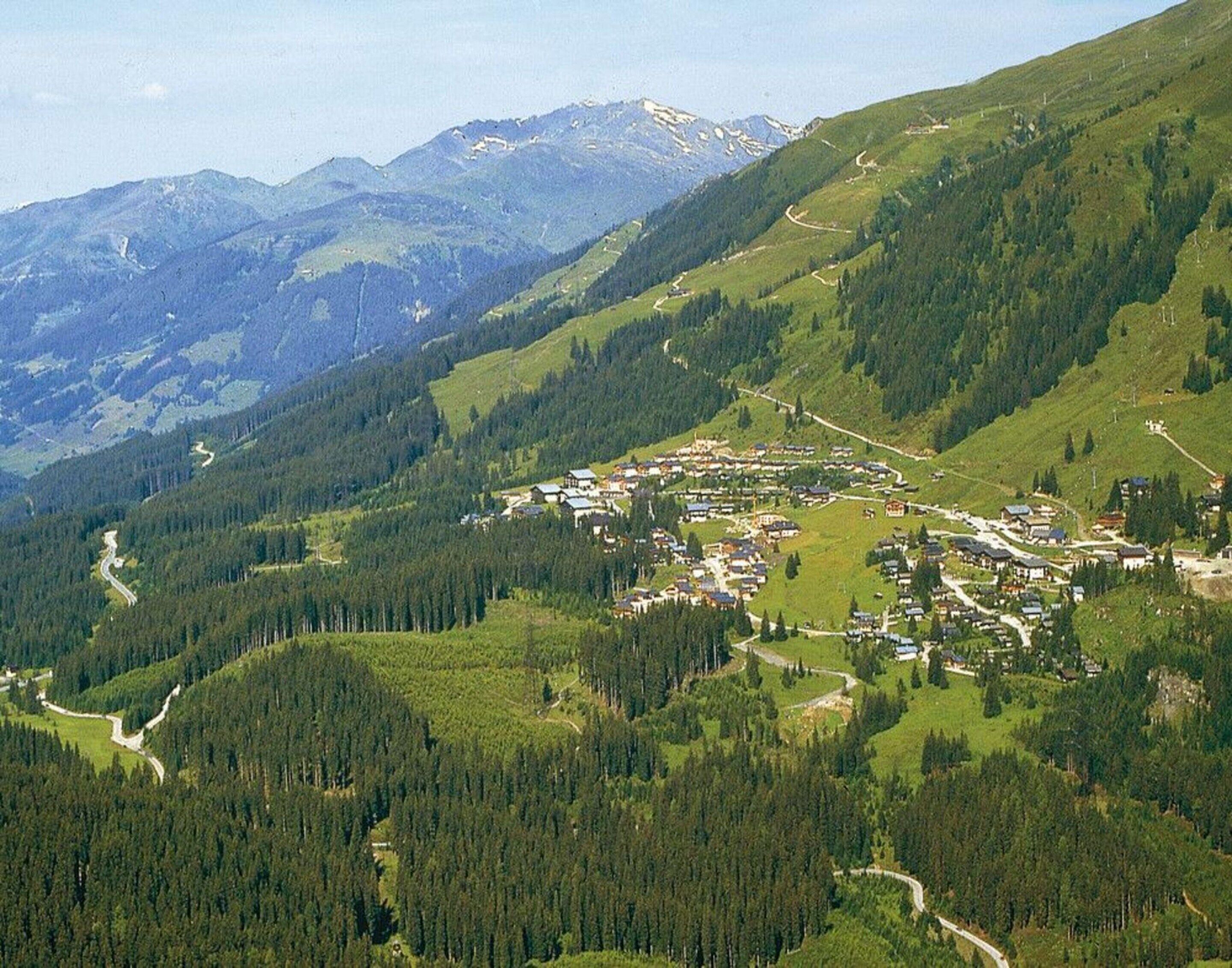 Un village de montagne, traversé par une route sinueuse, entouré de forêts denses et dominé, au loin, par des sommets enneigés.