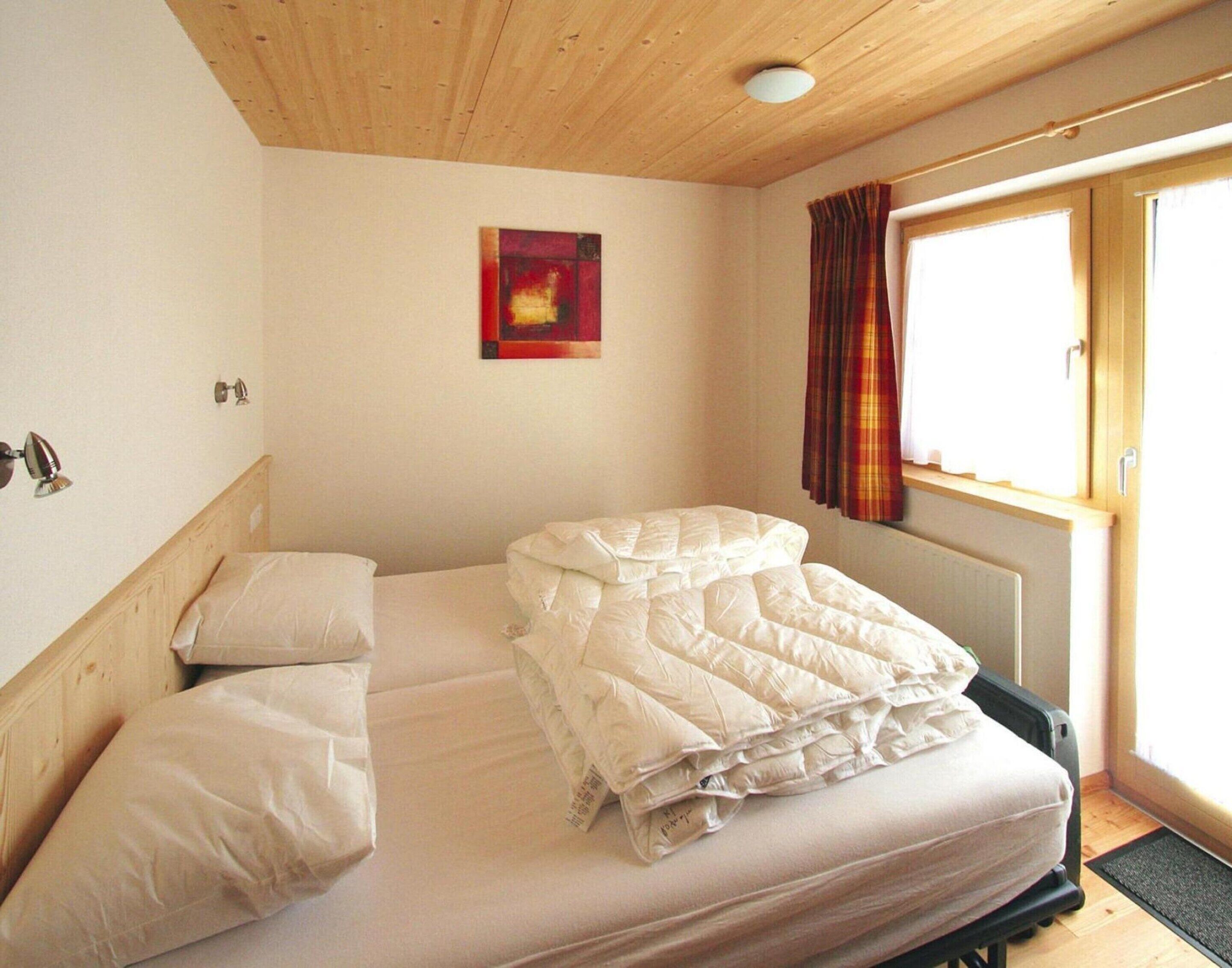 A single bed with a white mattress and pillows, a wooden headboard, a small painting on the wall, a window with curtains, and a radiator below.