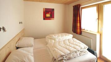 A single bed with a white mattress and pillows, a wooden headboard, a small painting on the wall, a window with curtains, and a radiator below.