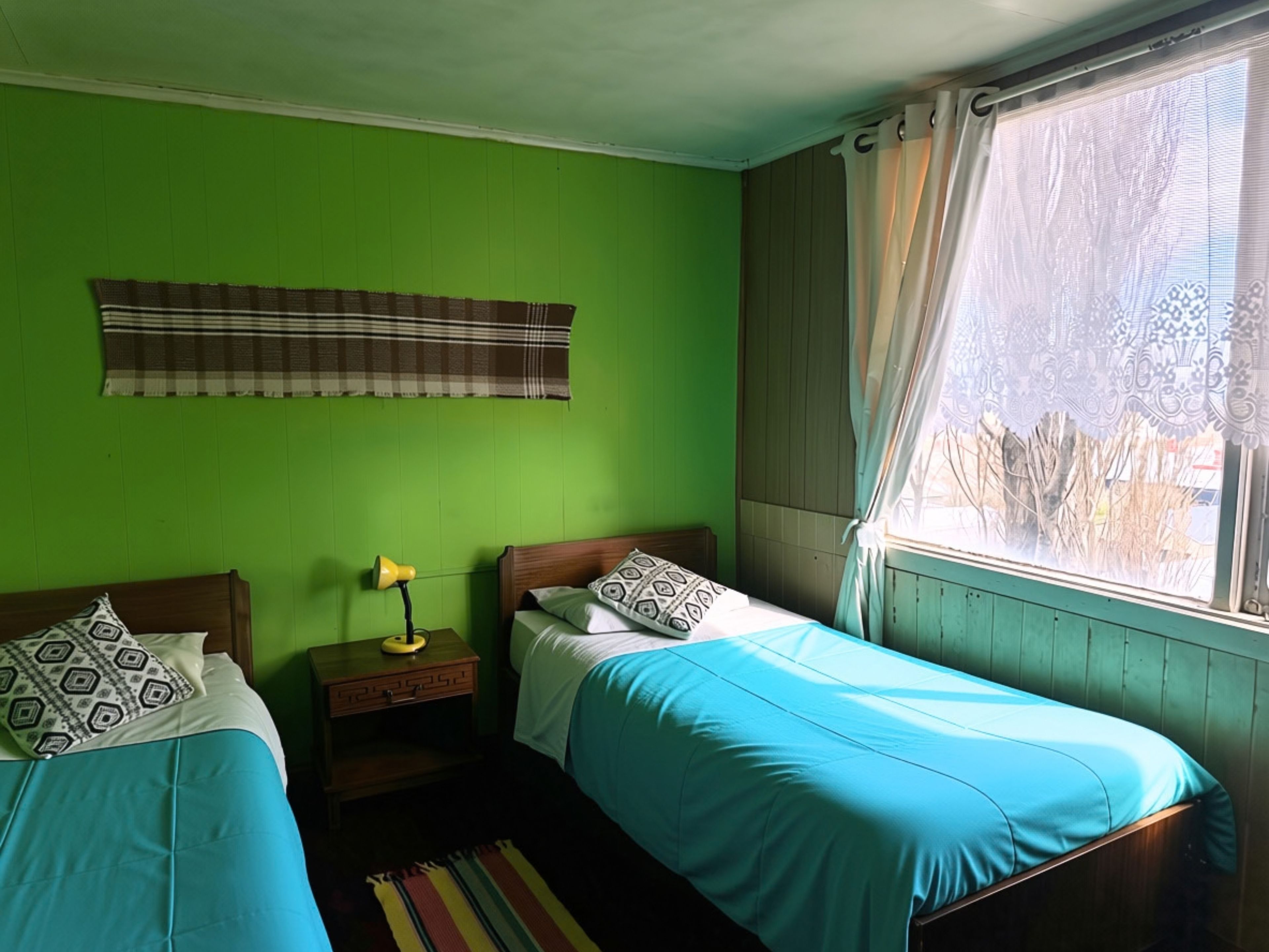 A room with two beds, a window with curtains, a lamp on a bedside table, and a striped rug.