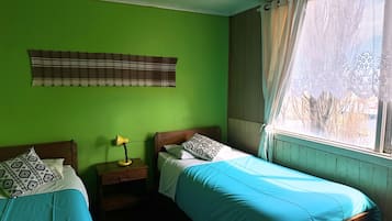 A room with two beds, a window with curtains, a lamp on a bedside table, and a striped rug.