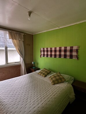 Ein Zimmer mit einem Bett, einer Wanddecke, einem Nachttisch mit Lampe und einem Fenster mit Vorhängen.