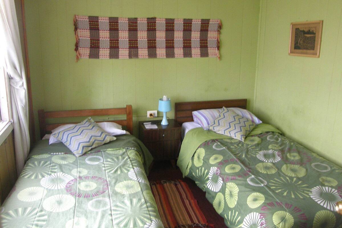 A room with two beds, each with patterned bedding, a blue lamp on a bedside table, and a checkered wall hanging.
