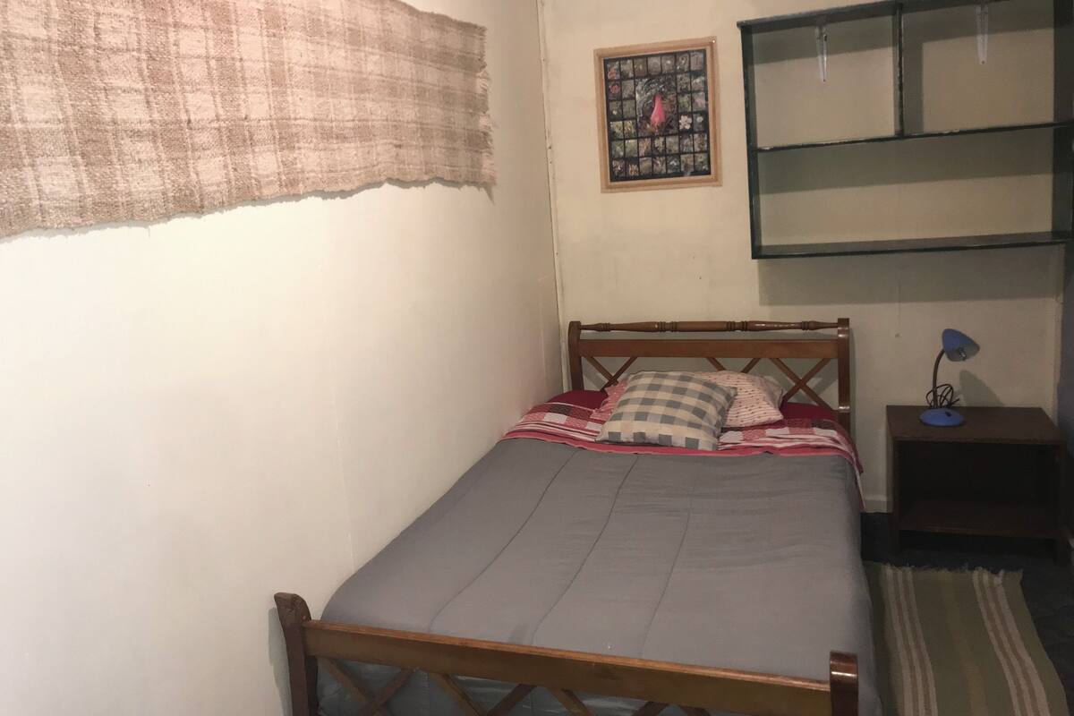 A small, simple bedroom with a single bed, a nightstand, a wall-mounted shelf, and a window with a curtain.