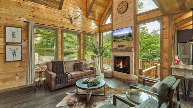 A cozy cabin interior with a fireplace, wooden walls, and large windows offering a view of the outdoors. - Views, Indoor Pool, Luxury Cabin, Bears (Gatlinburg)