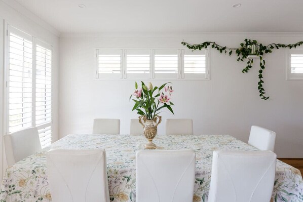 Une salle à manger avec une table dressée pour quatre personnes, des chaises blanches, une nappe à motifs floraux et un vase rempli de fleurs.