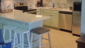 A kitchen with light green cabinets, a stainless steel refrigerator, a built-in oven, a microwave, and a coffee maker. There are two white bar stools with gray cushions. - Beautiful, Newly-Renovated Condo on the Altamaha River in Darien with BoatSlip (Darien)