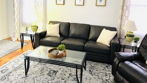 A living room with a black leather sofa, a glass coffee table, and a patterned rug. There are two framed botanical prints on the wall and two lamps on side tables.