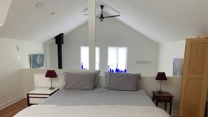 Ein Schlafzimmer mit schrägen Wänden, zwei Nachttischen mit Lampen, einem Bett mit weißer Bettdecke und einem Fenster mit blauen Vorhängen.
