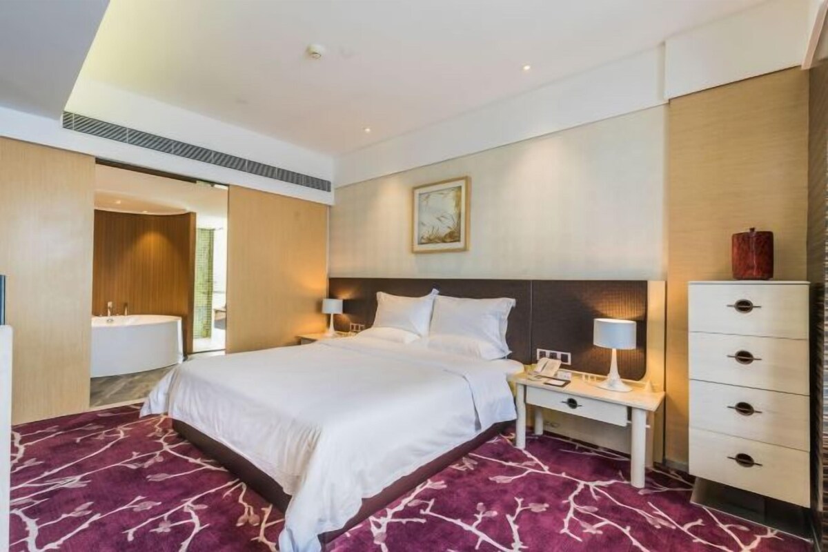 A hotel room with a large bed, a bedside table with a lamp, a nightstand, a dresser, and a bathroom visible through an open door.