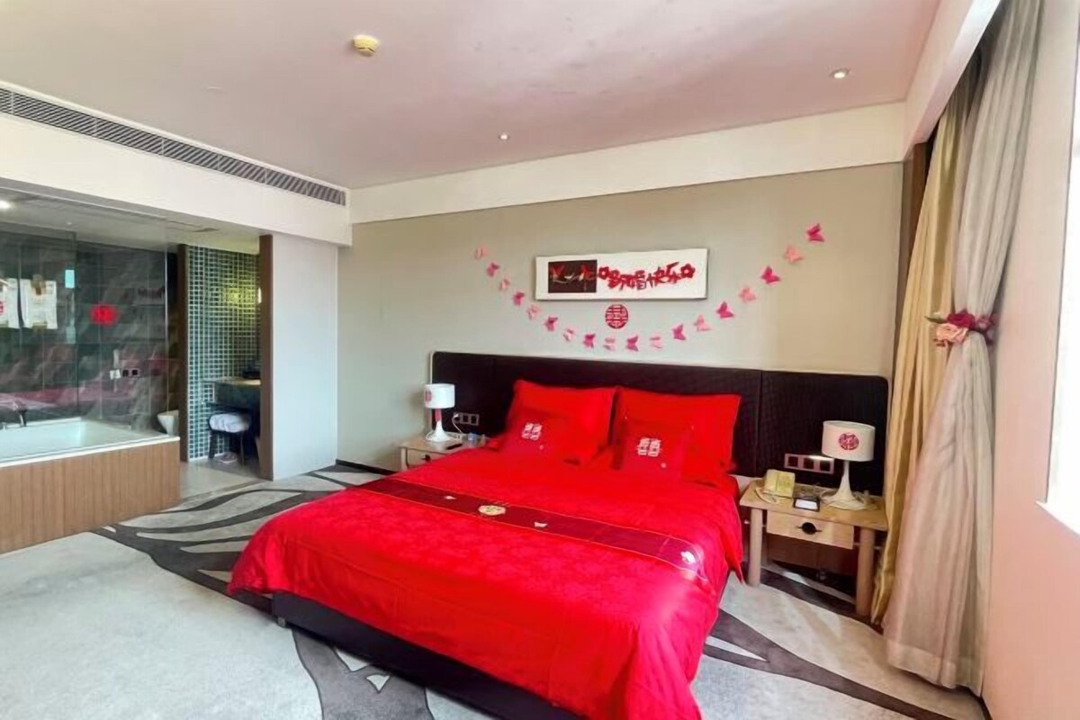 A modern hotel room with a large bed, a bedside table, a nightstand, a window with curtains, and a wall with a decorative item.