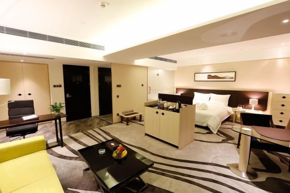 A hotel room with a bed, a desk, a chair, a sofa, a small table with fruit, and a decorative wall piece.