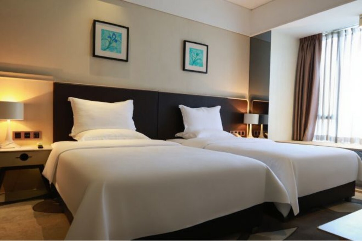 A hotel room with two beds, a nightstand, a lamp, and a window with curtains.